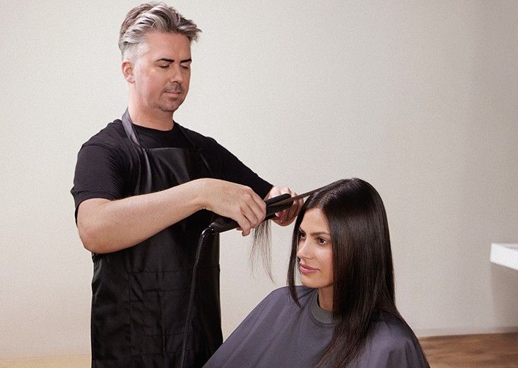 A stylist flat-ironing a strand of a seated client's long, straight dark hair, Kerasilk brand photo