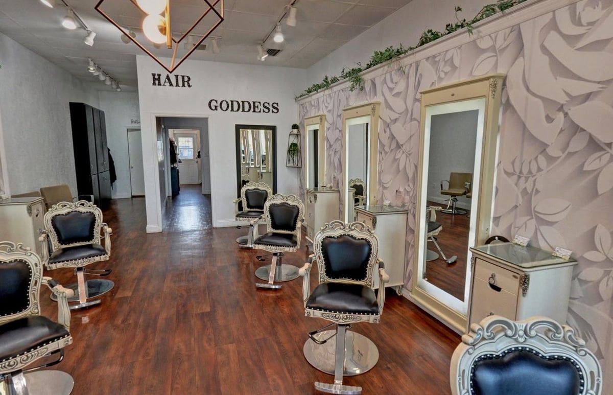 The styling floor at Hair Goddess Salon, Spa & Boutique: ornate white and black styling chairs, lighted mirrors along a floral wall, and the Hair Goddess name on the back wall
