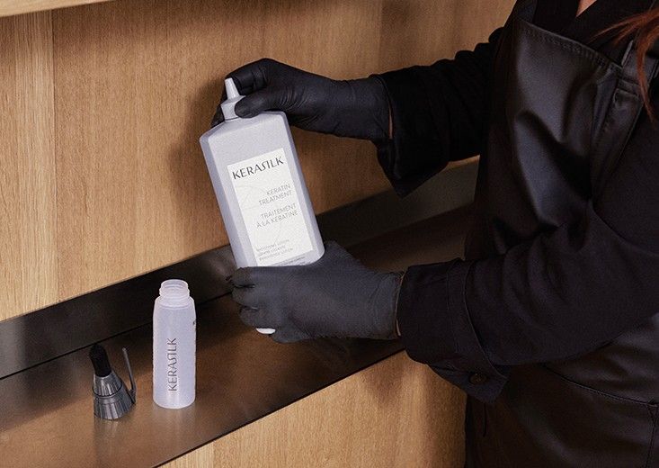 Gloved hands holding a Kerasilk Keratin Treatment bottle beside an empty applicator bottle on a salon shelf, Kerasilk brand photo